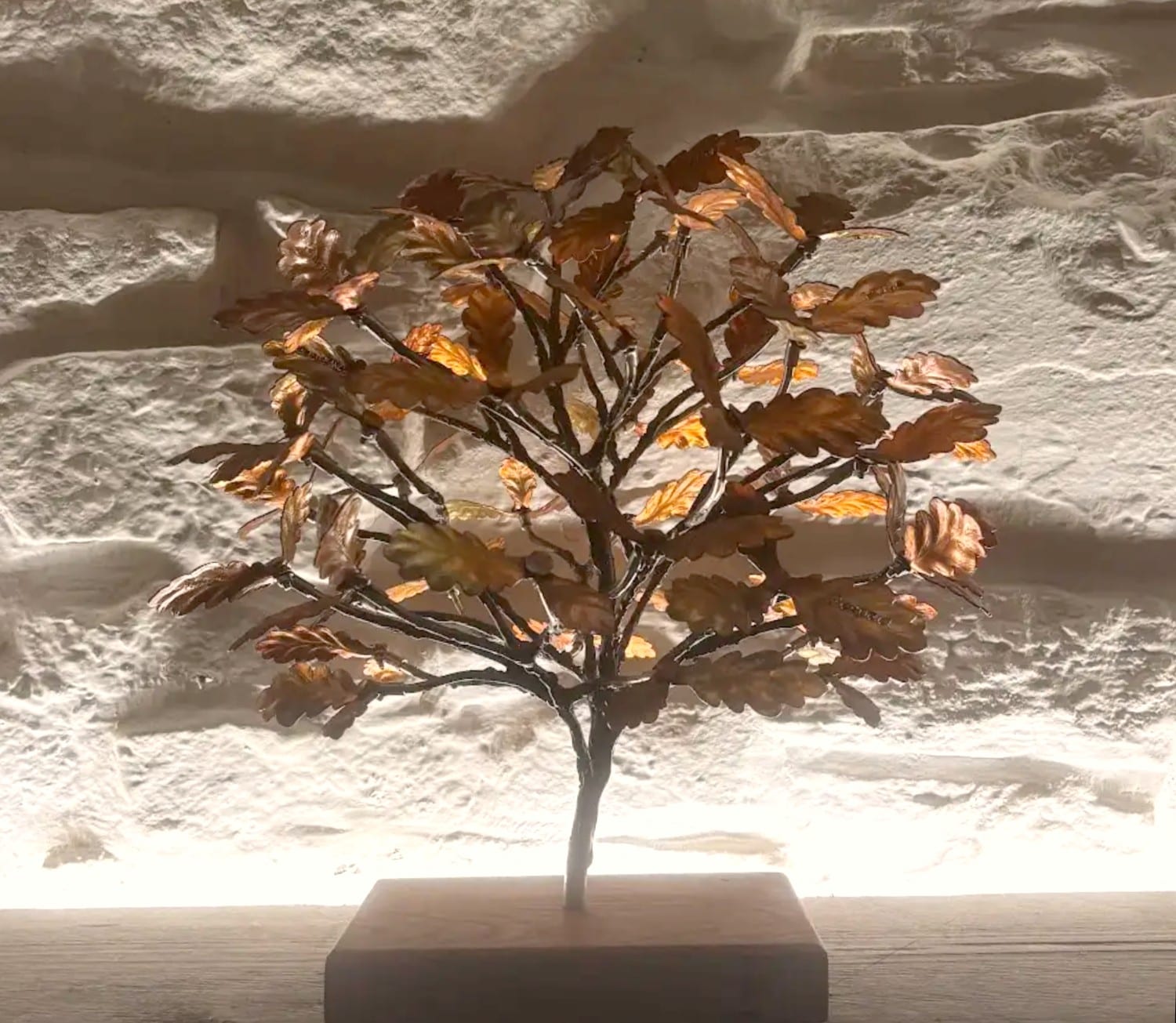 A small forged oak with copper leaves, lit from behind against a white stone wall